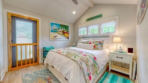 A bedroom with a bed, a nightstand, a window, a painting, and a door with a view.