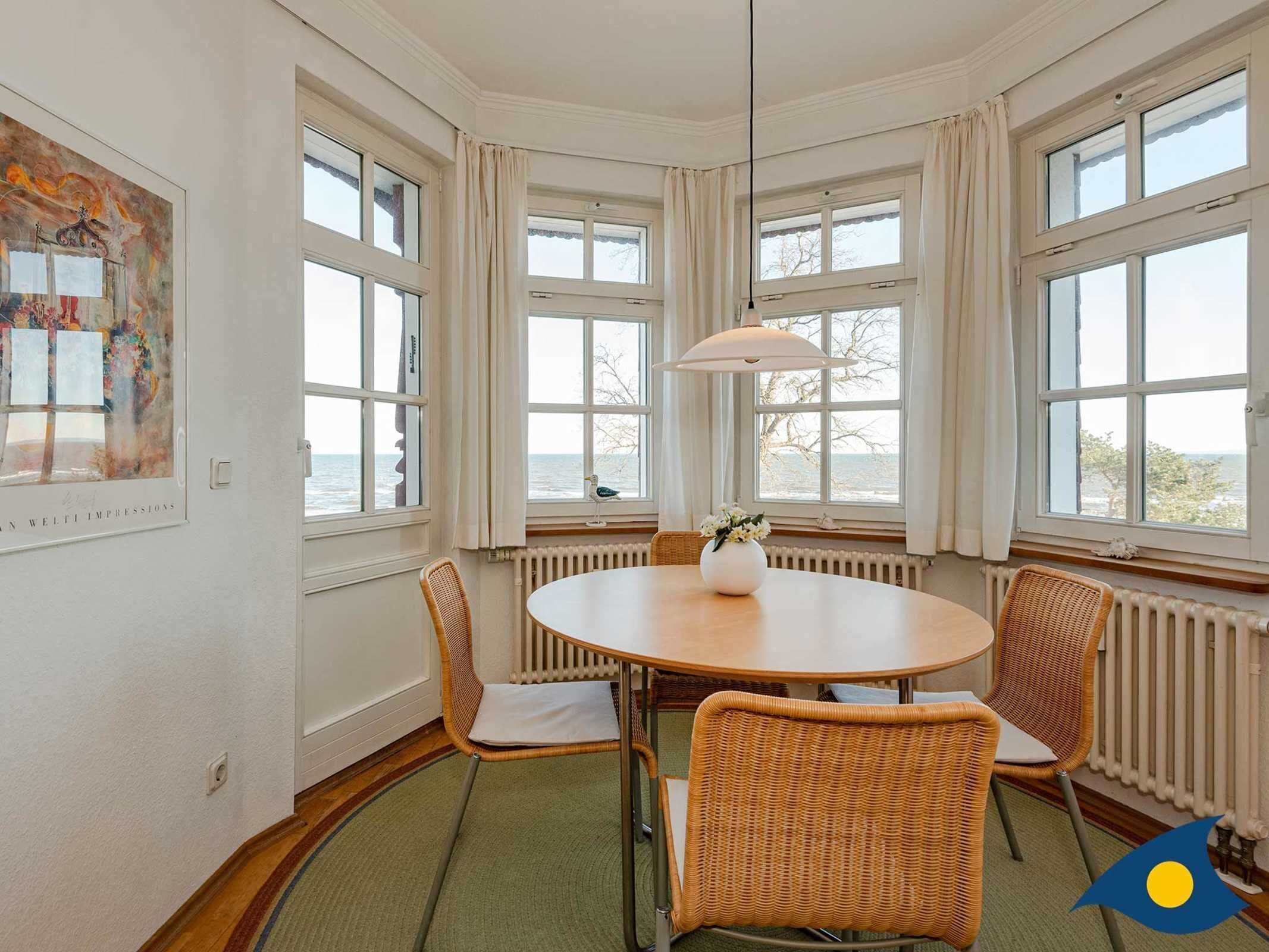 Une pièce avec une table ronde, des chaises en osier, de grandes fenêtres avec des rideaux, un radiateur et un tableau accroché au mur.