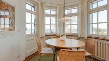 A room with a round table, wicker chairs, large windows with curtains, a radiator, and a painting on the wall.