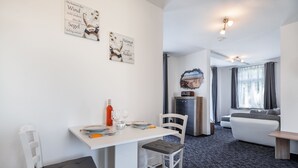 Ein modernes Wohnzimmer mit einer Couch, einem Esstisch gedeckt mit Tellern, Gläsern und einer Flasche. An den Wänden befinden sich Kunstwerke und eine Deckenleuchte.