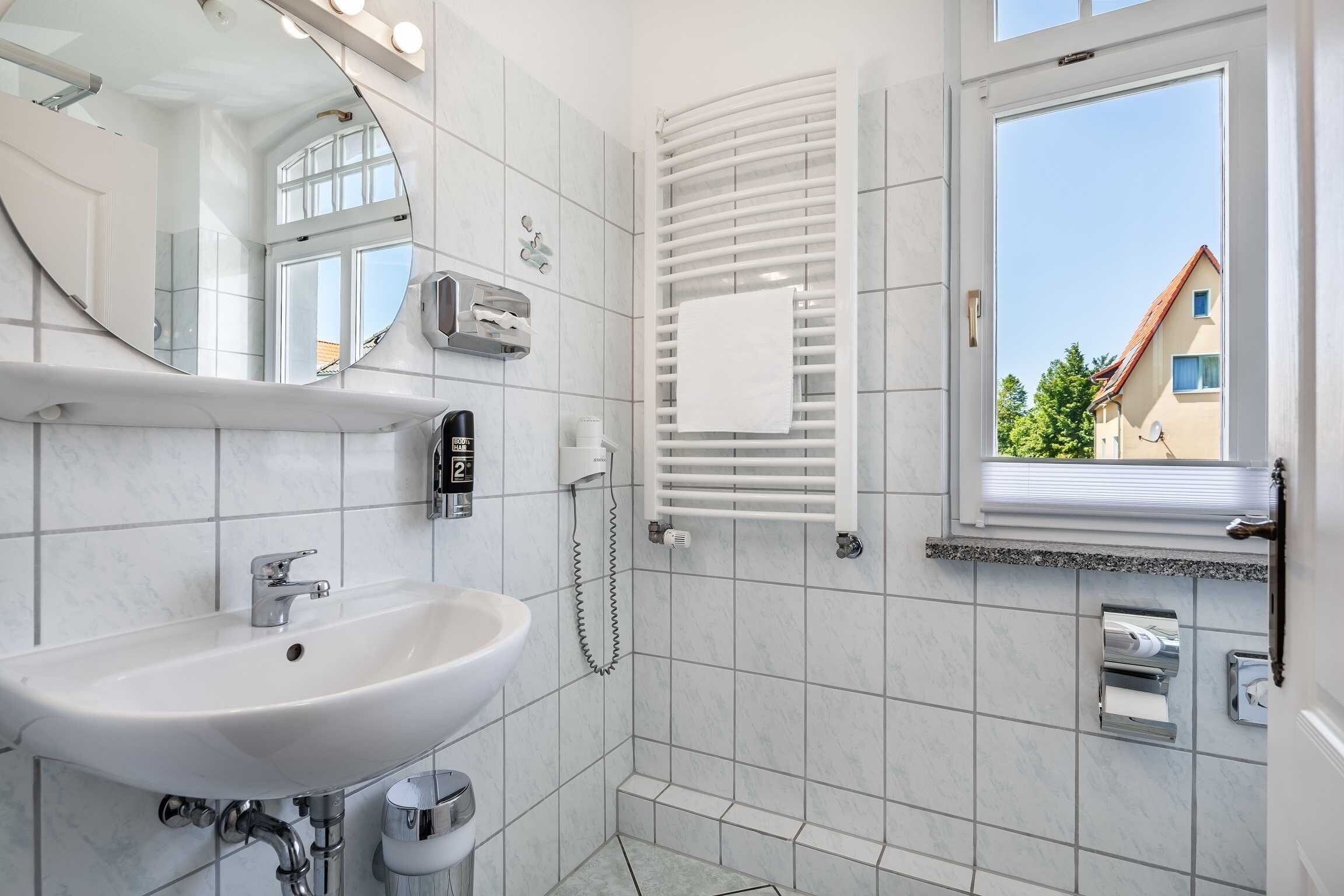 Ein Badezimmer mit einem weißen Waschbecken, einem Spiegel, einem Handtuchhalter und einem Fenster mit Blick auf Gebäude.