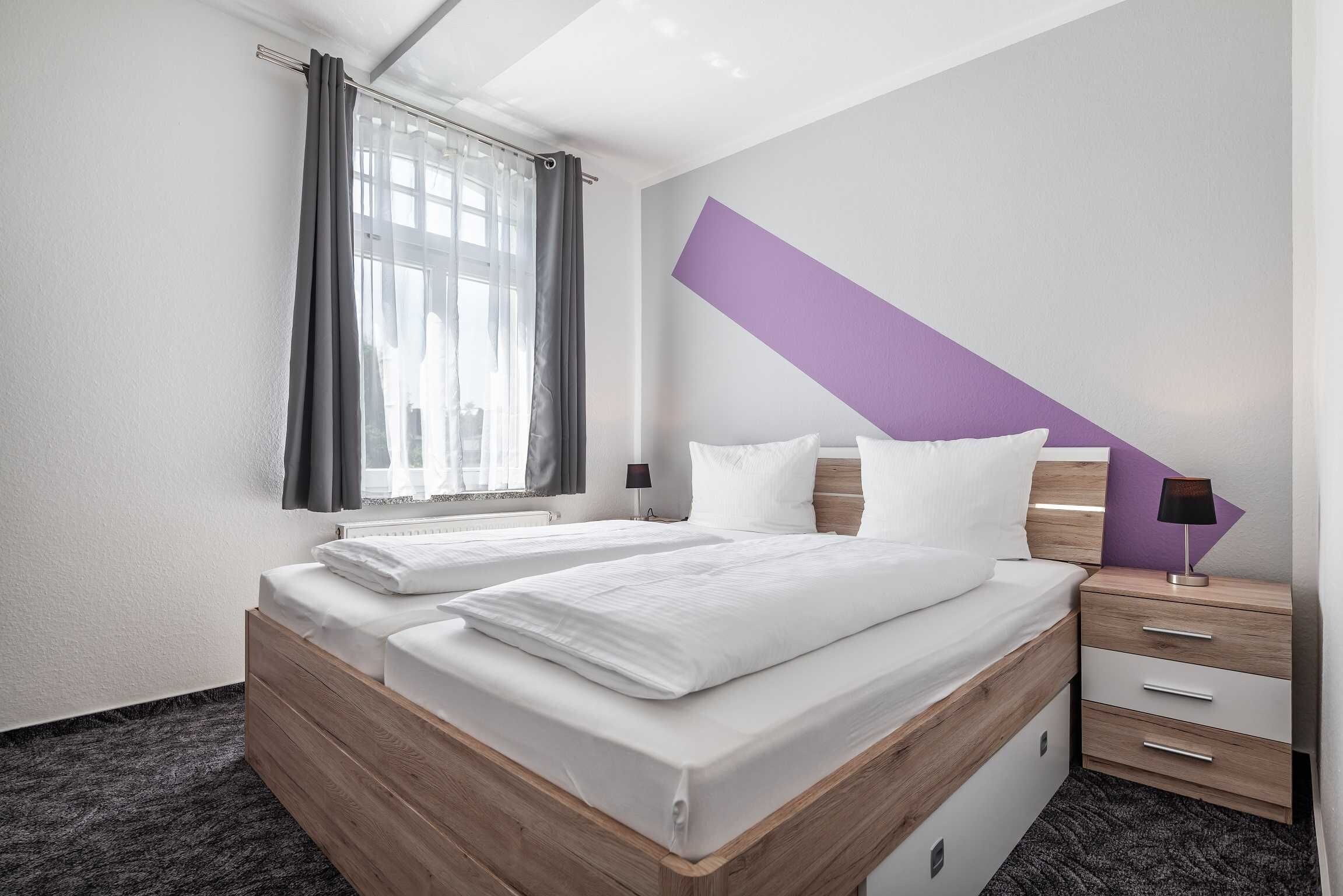 Une chambre d’hôtel avec deux lits, une fenêtre avec des rideaux, une table de chevet avec une lampe et un mur d’accent de couleur violette.