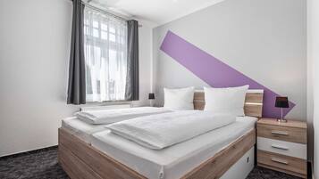 Een hotelkamer met twee bedden, een raam met gordijnen, een nachtkastje met een lamp en een paarse accentmuur.