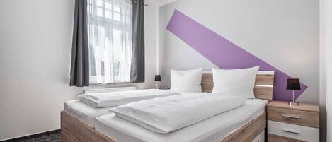 Une chambre d’hôtel avec deux lits, une fenêtre avec des rideaux, une table de chevet avec une lampe et un mur d’accent de couleur violette.