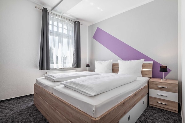Habitación de hotel con dos camas, una ventana con cortinas, una mesita de noche con lámpara y una pared de acento color púrpura.