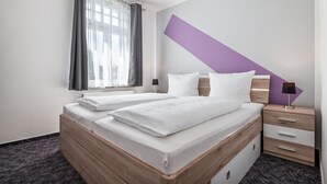 Ein modernes Hotelzimmer mit zwei Betten, einem Fenster mit Vorhängen, einem Nachttisch mit Lampe und einer Wand in Violett.