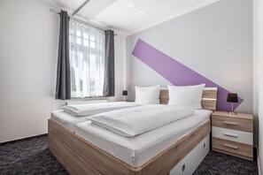 A modern hotel room with two beds, a window with curtains, a bedside table with a lamp, and a purple accent wall.