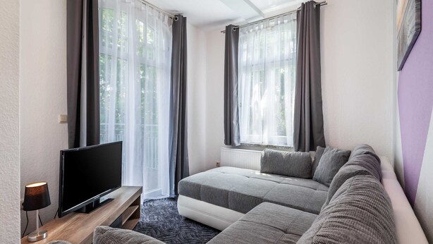 Ein modernes Wohnzimmer mit Flachbildfernseher, grauen Sofas und großen Fenstern mit transparenten Vorhängen.