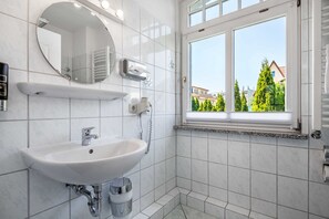 A modern bathroom with a white sink, a round mirror, and a window with a view of buildings and trees. - Apartment Rosenhof TW 113 - TW 113 (Kühlungsborn)