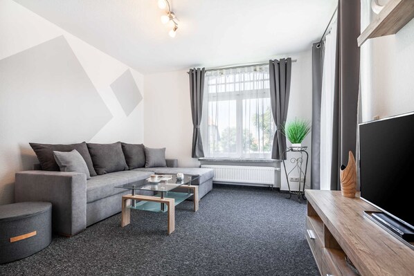 A modern living room with a grey sofa, a glass coffee table, and a flat-screen TV on a wooden stand.