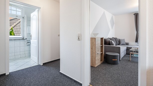 En moderne lejlighed med et opholdsområde, der indeholder en grå sofa, et træskab og en rund skammel. Der er et vindue med udsigt til en bygning med et tegltag.