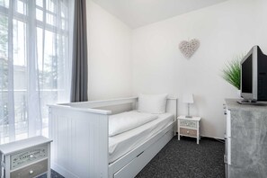 A small, white, single-bed room with a window, a heart-shaped wall decoration, a bedside table with a lamp, and a TV on a cabinet. - Apartment Rosenhof TW 113 - TW 113 (Kühlungsborn)