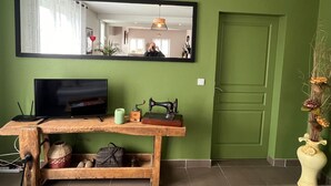 Une pièce avec un mur vert, un grand miroir, une console en bois sur laquelle se trouve un téléviseur, une machine à coudre et un vase rempli de fleurs séchées.