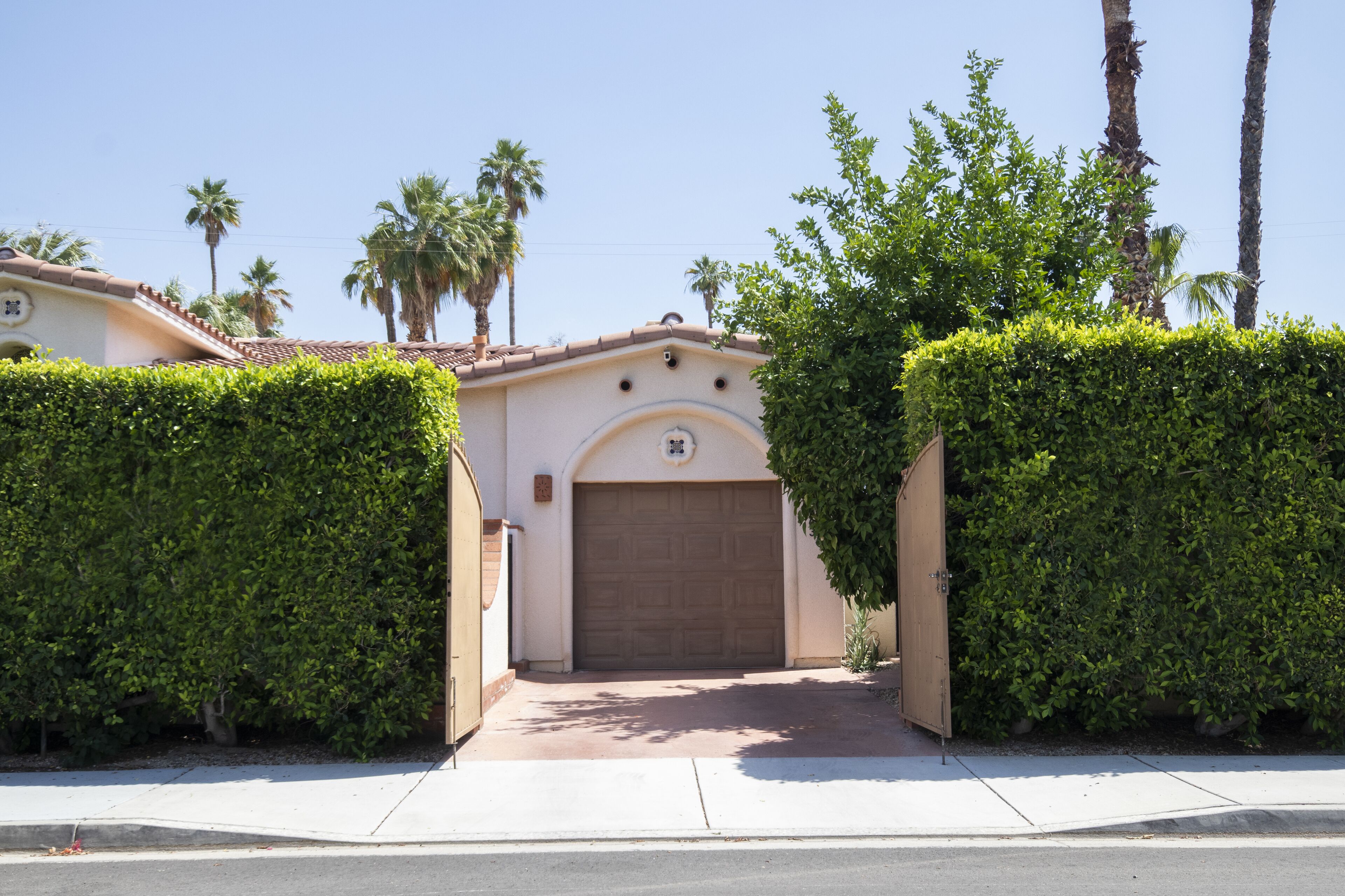 A residential house with a brown garage door, surrounded by well-maintained hedges and palm trees.