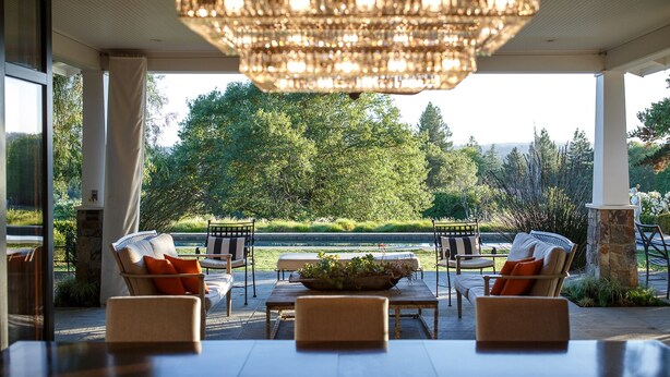 A spacious outdoor seating area with a large table and chairs, a decorative chandelier, and a view of a lush garden and trees.