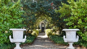 A gravel pathway flanked by neatly trimmed hedges and two white urns with classical designs.