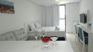 Ett modernt hotellrum med en säng, en soffa, ett matbord med glas och en skål med frukt, en tv och en tavla på väggen.