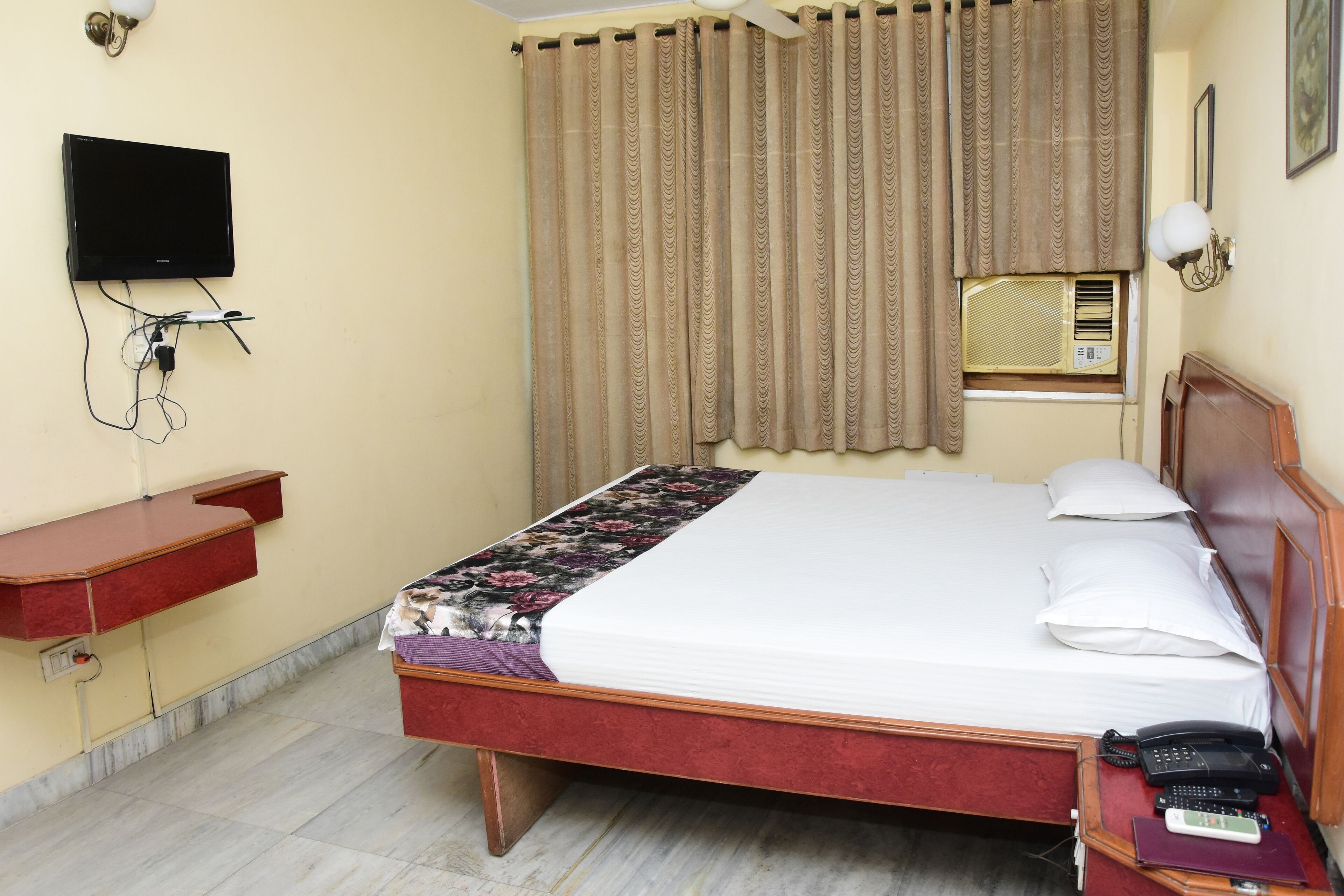 A hotel room with a bed, bedside table, TV, air conditioning unit, and curtains.