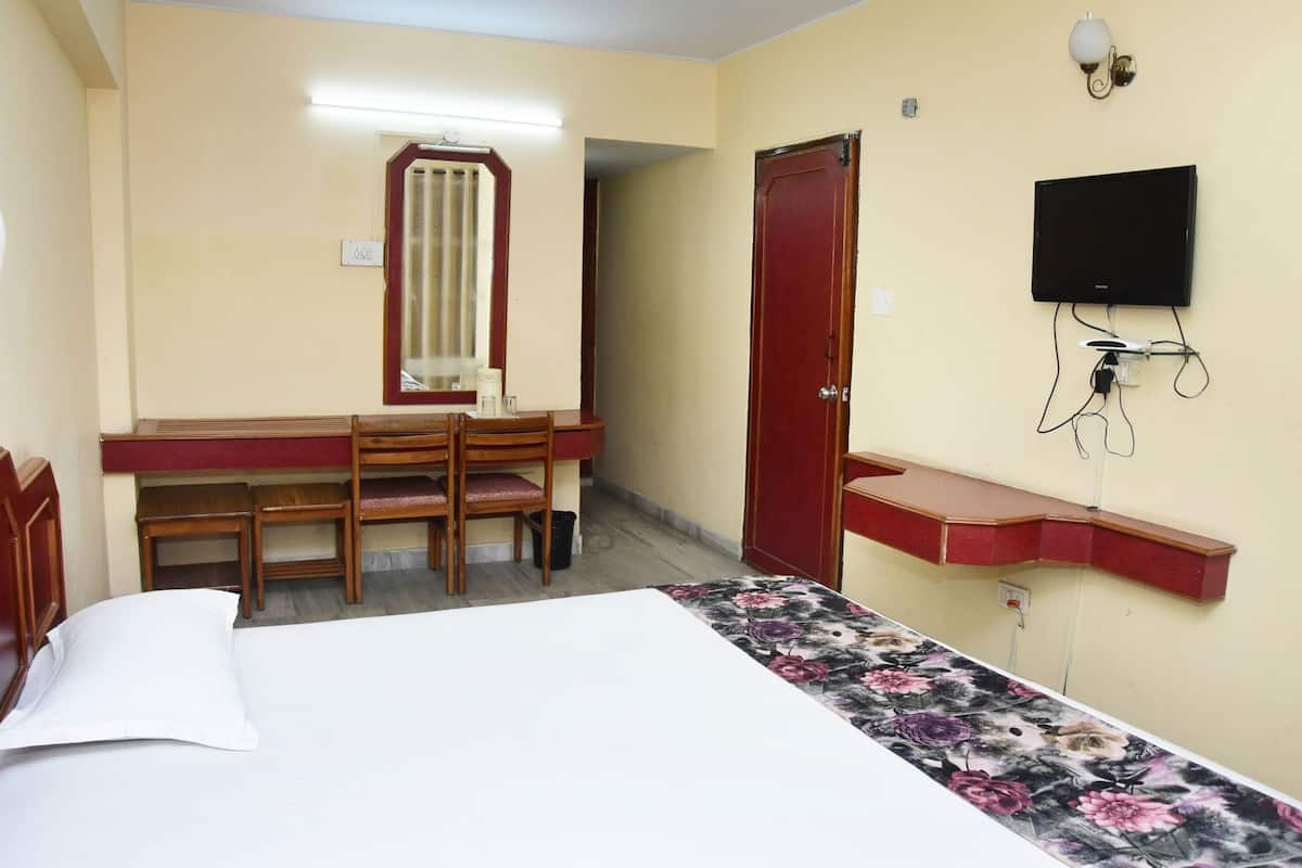 Habitación de hotel con cama, escritorio con sillas, espejo, puerta roja, televisor montado en la pared y una pequeña estantería.