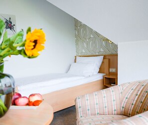 A hotel room with a bed, a wooden nightstand, a patterned headboard, a small table with fruit, and a sofa.