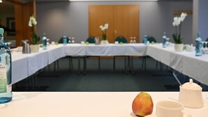 Ein Konferenzraum mit einem langen Tisch, Stühlen und einer weißen Tischdecke. Auf dem Tisch stehen Wasserflaschen, Obst und eine Tasse.