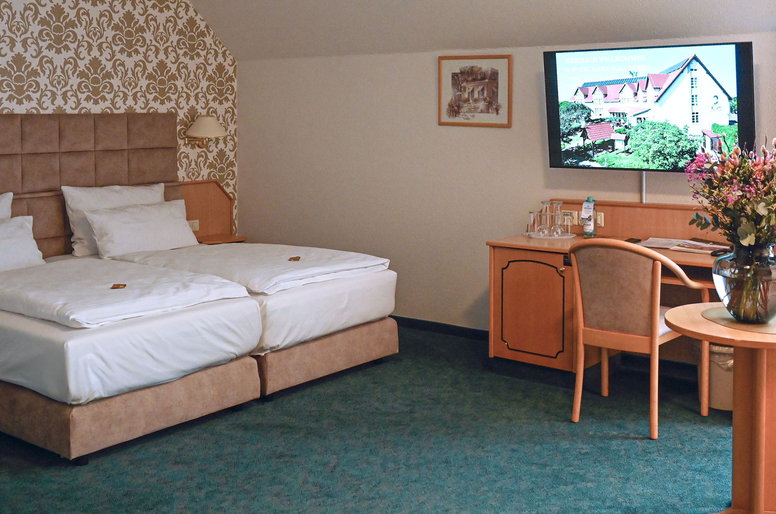 Et hotelværelse med en dobbeltseng, et skrivebord med stol, et fjernsyn og et lille bord med blomster.