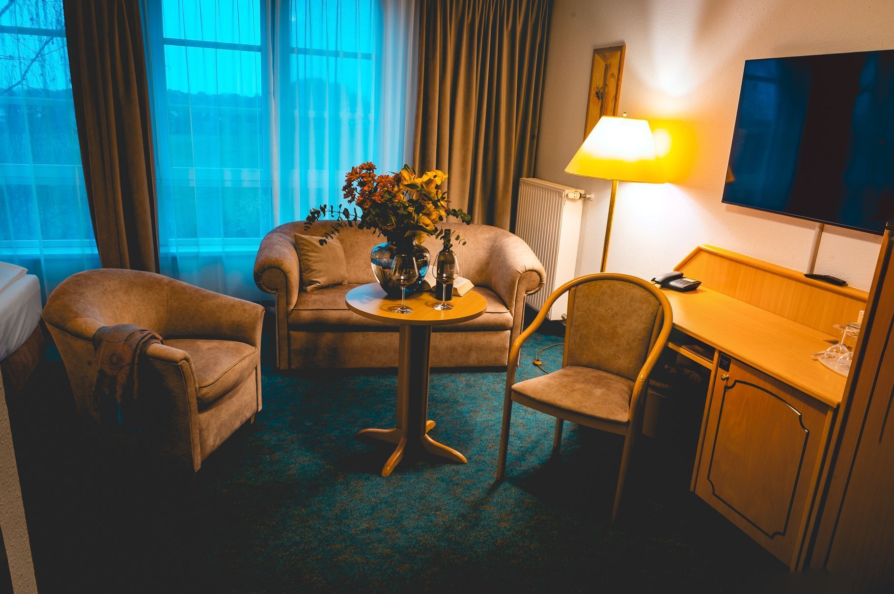 Ein Hotelzimmer mit einer Couch, zwei Sesseln, einem kleinen Tisch mit einer Vase und Weingläsern, einem Schreibtisch mit einer Lampe und einem Fernseher.