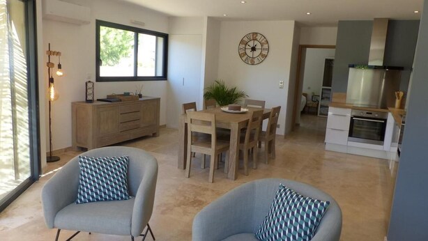 A modern kitchen with a built-in oven, a dining area with a wooden table and chairs, and two grey armchairs with patterned cushions.