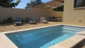 A rectangular swimming pool with a tiled edge, surrounded by a gravel area. There are lounge chairs and a table with chairs in the background.