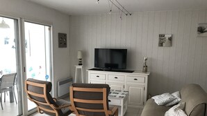 A living room with a TV, two wooden chairs, a sofa, a small table, and a sliding glass door leading to an outdoor area. - House for rent 200 meters from the beach, close to shops. (Saint-Jean-de-Monts)