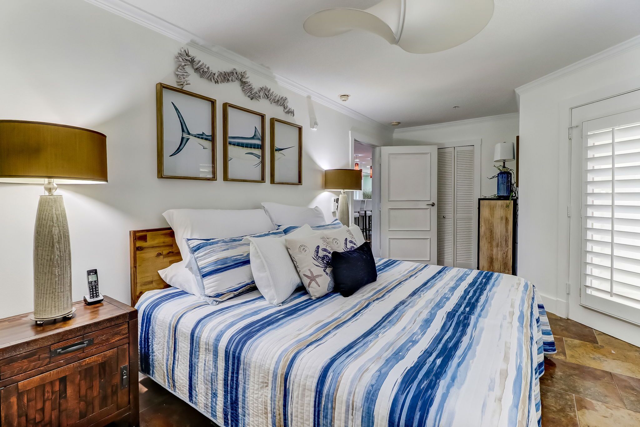 A bedroom with a bed, bedside tables, and a nightstand. There are framed pictures on the wall, a ceiling light, and a window with blinds.