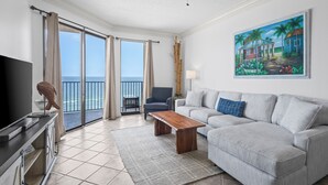 A spacious living room with a large sofa, a wooden coffee table, and a television on a cabinet. There is a painting of a tropical scene on the wall, and a balcony with a view of the ocean. - BEAUTIFUL BEACHFRONT CONDO, FAMILY AMENITIES, GREAT GULF VIEWS! (Orange Beach)