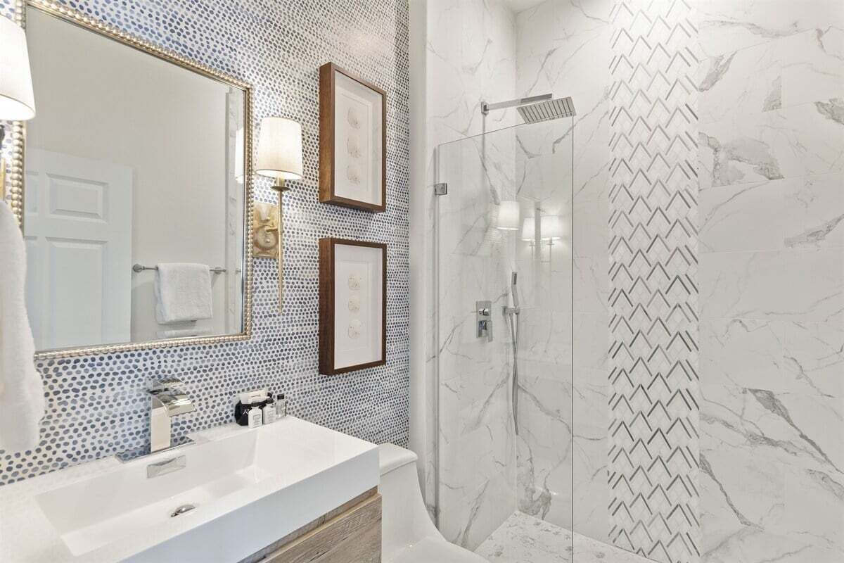Een badkamer met een witte wastafel, een grote spiegel, afbeeldingen in lijsten en een douche met een behangpatroon.