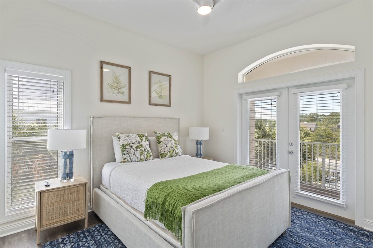A bedroom with a bed, two bedside lamps, a nightstand, a window with blinds, and a door with a balcony.