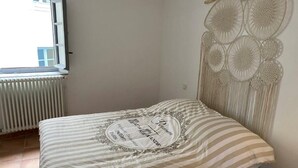 A single bed with a striped bedspread and a decorative pillow, a radiator, and a macrame wall hanging in a room with plain walls. - Mansion overlooking the ancient Roman arena (Béziers)