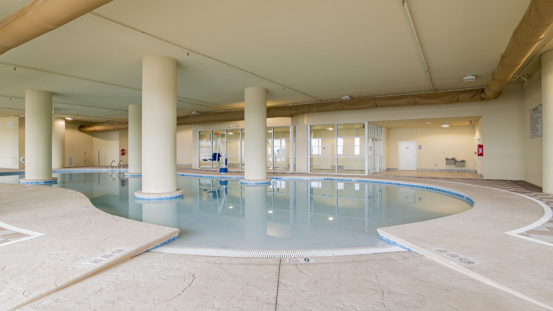 Piscina interna com água cristalina, cercada por colunas e paredes bege, e piso ladrilhado.