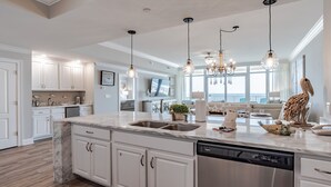A modern kitchen with white cabinetry, a marble countertop, and a dishwasher. There is a dining area with a view of the ocean. - New Complex!  Lazy River/Slide, Beachfront!  5th Floor (Orange Beach)