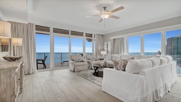 A spacious living room with a large white sofa set, a coffee table, and a dining area with a view of the ocean. - New Complex!  Lazy River/Slide, Beachfront!  4BR, Sleeps 14! (Orange Beach)