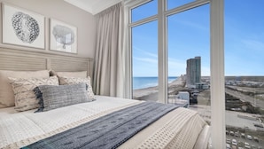 A bedroom with a large bed, a view of the beach, and two framed botanical prints on the wall. - New Complex!  Lazy River/Slide, Beachfront!  4BR, Sleeps 14! (Orange Beach)