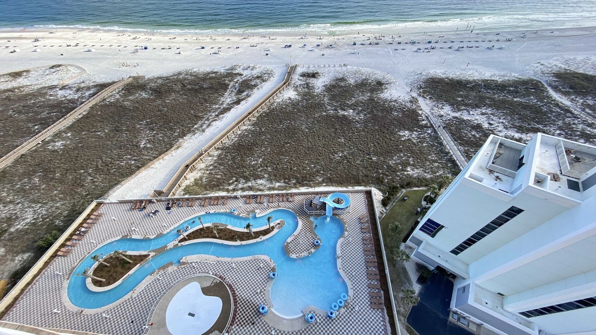 Eine Vogelperspektive zeigt einen Urlaubsort mit einem großen Poolbereich, einem Strand und einem Hochhaus.