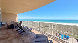 A balcony with a view of the beach, a table with chairs, and a pool area.