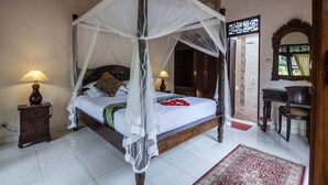 Un lit à baldaquin à quatre poteaux, recouvert d’une courtepointe blanche et agrémenté de fleurs rouges, flanqué de deux lampes de chevet, dans une pièce dotée d’un plancher en bois et d’un tapis à motifs rouges et blancs.