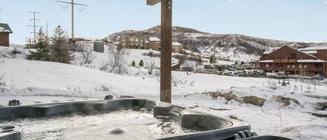 A hot tub area with a wooden structure overhead, a snowy landscape, and a building in the background.