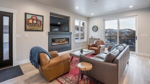 A modern living room with a fireplace, leather sofas, a coffee table, and large windows offering a view of the outdoors. - 845 Mill Run Court 5 Bedroom Home by RedAwning (Steamboat Springs)