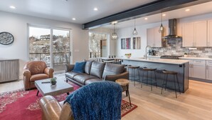 A modern living room with a sofa, armchairs, and a coffee table. There is a kitchen area with a large island and bar stools. - 845 Mill Run Court 5 Bedroom Home by RedAwning (Steamboat Springs)