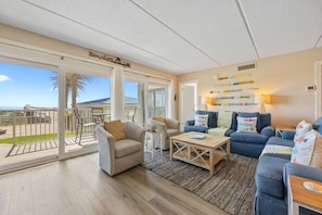 A spacious living area with a large sliding glass door to a balcony, a blue sofa set, a wooden coffee table, and a wall-mounted lamp.
