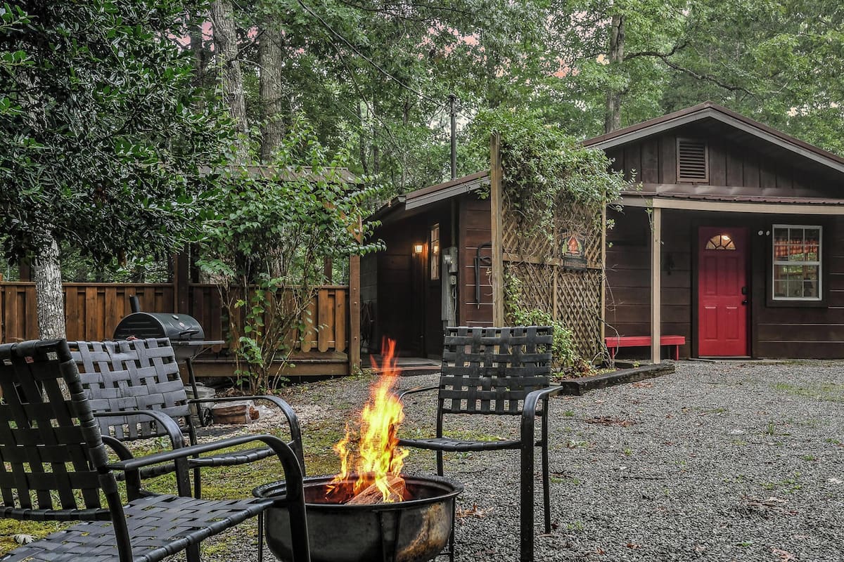 A rustic cabin with a red door, a fire pit in the center, and a wooden deck.