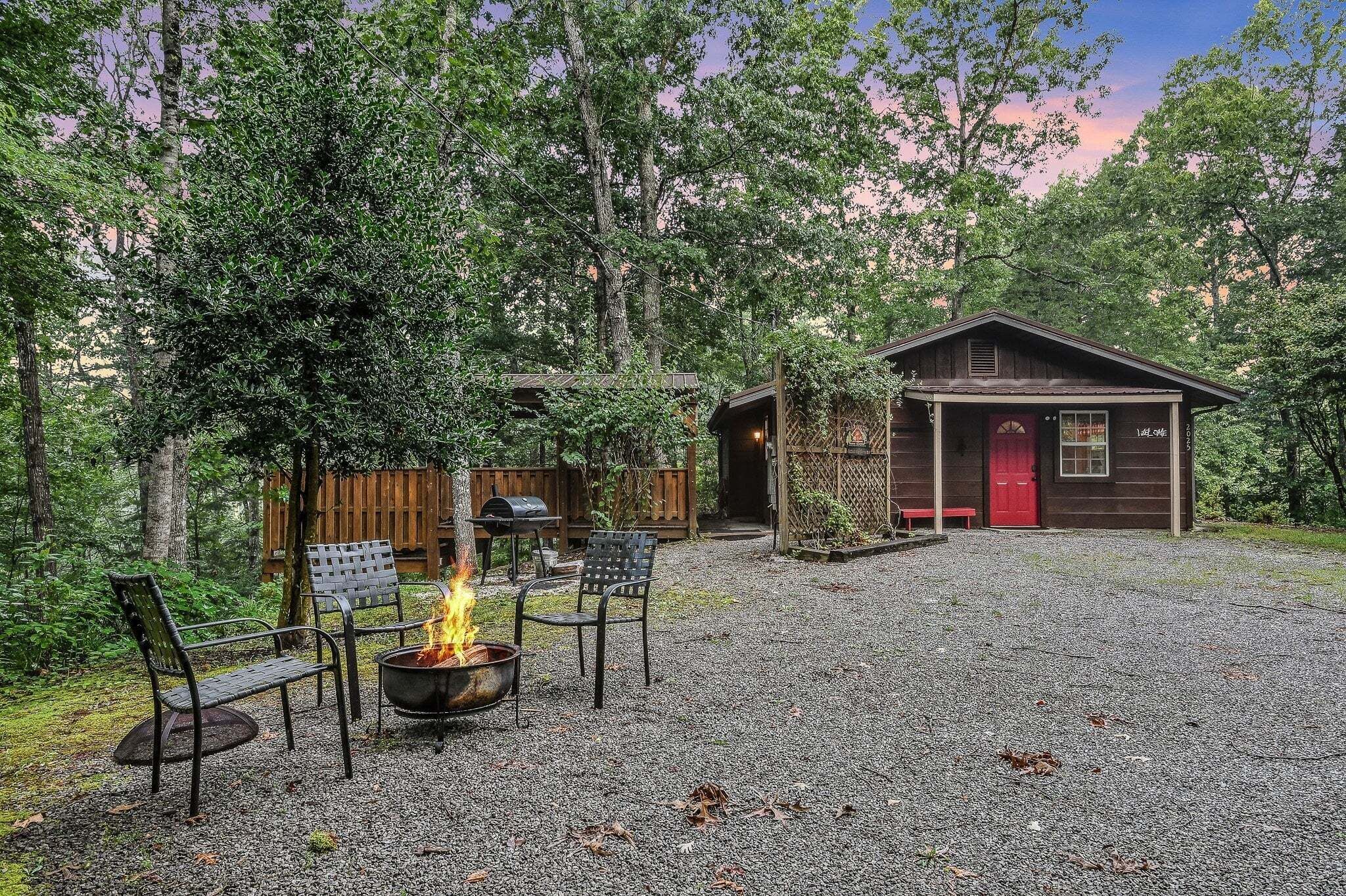 A rustic cabin with a gravel driveway, surrounded by trees. There is a fire pit with chairs and a bench.