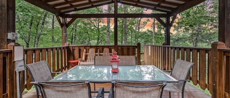 A covered outdoor patio with a glass table and chairs, surrounded by wooden railings and trees.