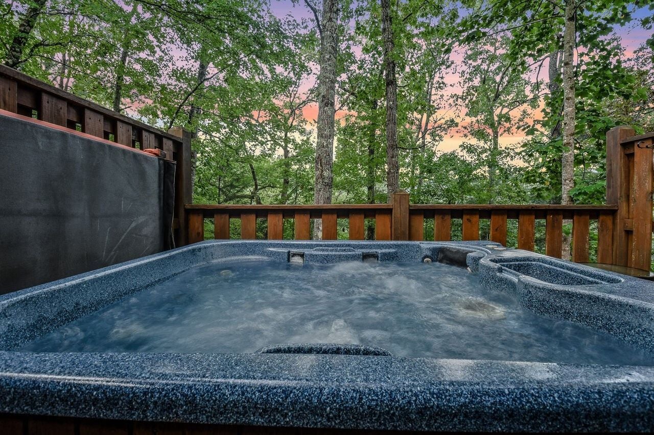A hot tub surrounded by a wooden deck with a view of trees and a sunset.